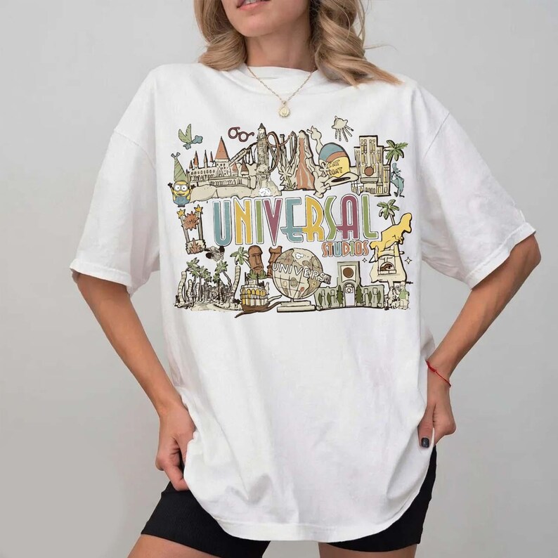 May include: White t-shirt with a colourful graphic of Universal Studios attractions and the text "UNIVERSAL STUDIOS". The design includes cartoon illustrations of various theme park elements. The shirt has short sleeves.