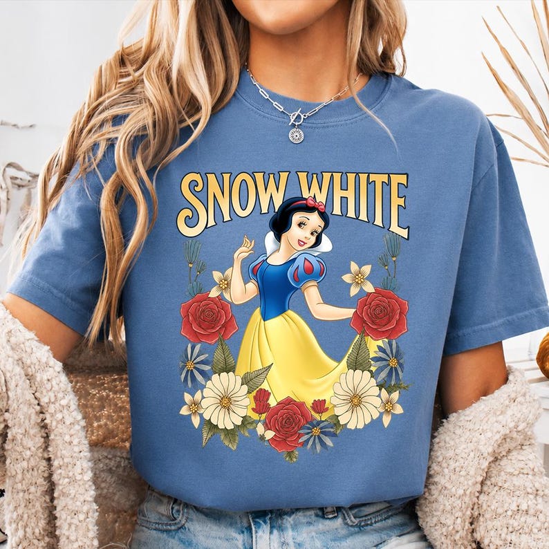 Puede incluir: Camiseta azul con un dise&ntilde;o de Blancanieves con un vestido amarillo y una parte superior azul, rodeada de rosas rojas y flores blancas. Las palabras "SNOW WHITE" est&aacute;n arqueadas sobre el personaje en letras amarillas.