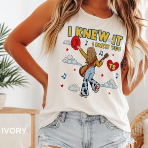 May include: An ivory sleeveless tank top featuring a graphic illustration of a person with long hair wearing a yellow plaid shirt and cow-print bell-bottom jeans, holding a guitar and a red hat, with the text I KNEW IT I KNEW YOU and a red heart.