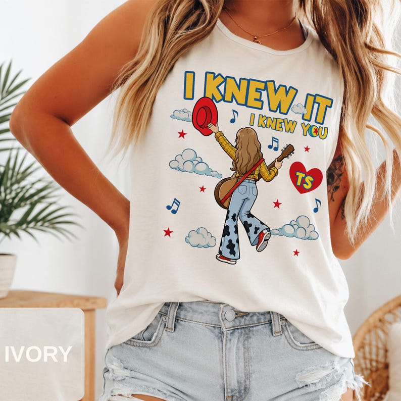 May include: An ivory sleeveless tank top featuring a graphic illustration of a person with long hair wearing a yellow plaid shirt and cow-print bell-bottom jeans, holding a guitar and a red hat, with the text I KNEW IT I KNEW YOU and a red heart.