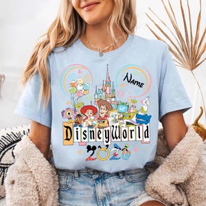 Peut inclure: T-shirt bleu clair avec un motif Disney World color&eacute;. Le motif comprend des personnages Disney, un ch&acirc;teau, des ballons et le texte "Disneyworld 2023". Un espace est disponible pour ajouter un nom.
