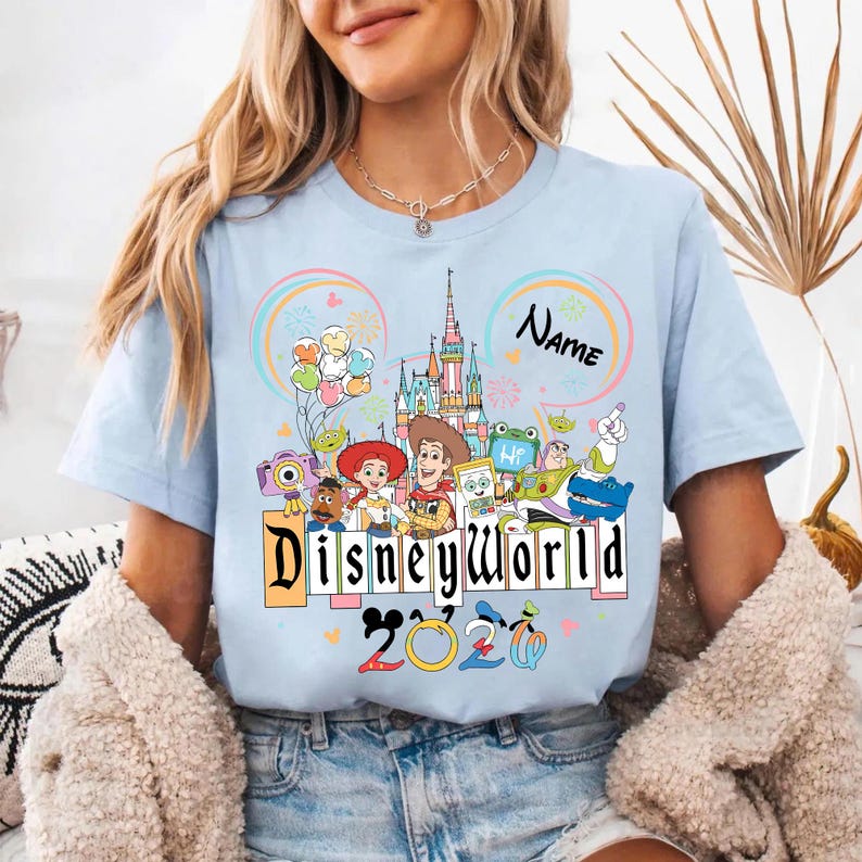 Peut inclure: T-shirt bleu clair avec un motif Disney World color&eacute;. Le motif comprend des personnages Disney, un ch&acirc;teau, des ballons et le texte "Disneyworld 2023". Un espace est disponible pour ajouter un nom.