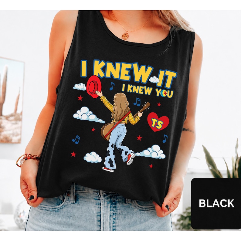 May include: A black sleeveless tank top featuring a graphic illustration of a person with long hair wearing a yellow plaid shirt and cow-print pants, holding a guitar and a red hat. The text reads I KNEW IT I KNEW YOU with a red heart labeled TS.