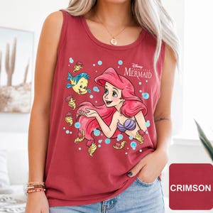 Peut inclure: Un d&eacute;bardeur sans manches cramoisi avec une impression graphique d'Ariel de La Petite Sir&egrave;ne, ses longs cheveux rouges, un haut coquillage violet et le poisson Polochon, entour&eacute;s de petits hippocampes jaunes et de bulles bleu clair.