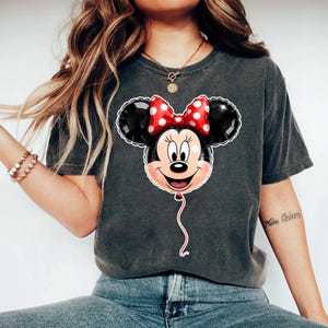 May include: A charcoal gray short-sleeve t-shirt featuring a graphic print of a Minnie Mouse-shaped balloon. The balloon is black with a red and white polka-dot bow, and it includes a white string hanging from the bottom of the character's chin.