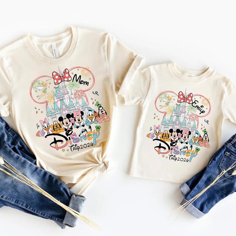 May include: Two cream-colored t-shirts with a Disney theme. One shirt says "Mom" and the other says "Emily". Both shirts feature a castle, Disney characters, and the text "Disney Trip 2026".