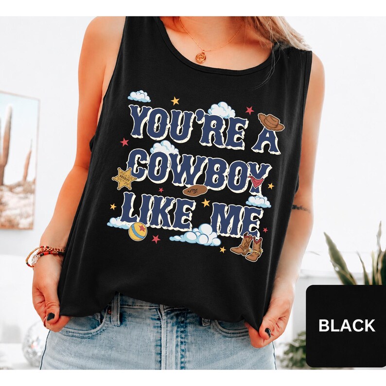 May include: A black sleeveless tank top featuring the text "YOU'RE A COWBOY LIKE ME" in a bold blue font. The design includes graphic illustrations of a brown cowboy hat, a sheriff star badge, a red bandana, a colourful ball, and brown boots.