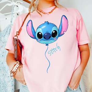 May include: A light pink short-sleeve t-shirt featuring a large graphic of a blue Stitch character designed as a balloon with pink inner ears and large black eyes. Below the balloon, the word Stitch is written in a light blue cursive script.