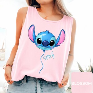 May include: A light pink sleeveless tank top featuring a graphic of a blue character shaped like a balloon with large black eyes and pink inner ears. Below the balloon, the word Stitch is written in a light blue cursive script font.