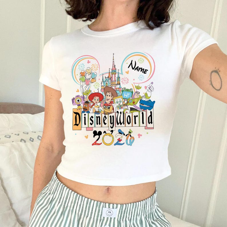 Peut inclure: Haut court blanc avec un motif color&eacute; de Disney World. Le motif comprend des personnages de dessins anim&eacute;s, un ch&acirc;teau et le texte "Disneyworld 2020". Le mot "Name" est dans un cercle.