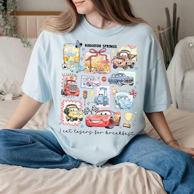 May include: Light blue t-shirt featuring a collage of cartoon cars and the text "Radiator Springs." The design includes various car characters and the phrase "I eat losers for breakfast" in a playful font. The shirt is a casual wear item.
