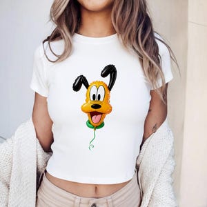 May include: A white short-sleeved t-shirt featuring a graphic print of a yellow and black Pluto character balloon. The balloon has a shiny, inflated appearance with a green knot at the base and a thin green string dangling downwards.