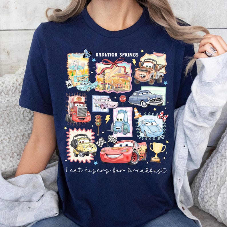 May include: Navy blue t-shirt featuring a colorful graphic of various cartoon cars and the text "Radiator Springs". The design includes a red race car, a yellow car, and a trophy. The phrase "I eat losers for breakfast" is also printed on the shirt.
