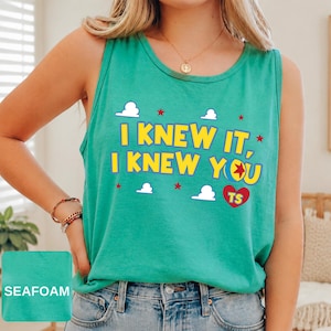 May include: A seafoam green sleeveless tank top featuring the yellow text I KNEW IT, I KNEW YOU with a colourful ball graphic, a red heart with the letters TS, and white cloud and red star accents printed on the front of the garment.