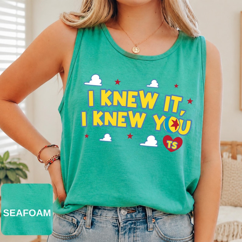 May include: A seafoam green sleeveless tank top featuring the yellow text I KNEW IT, I KNEW YOU with a colourful ball graphic, a red heart with the letters TS, and white cloud and red star accents printed on the front of the garment.