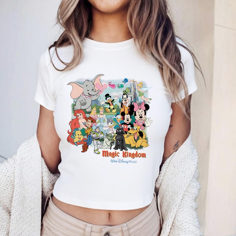 K&ouml;nnte beinhalten: Wei&szlig;es Crop-Top mit einer farbenfrohen Grafik von Disney-Figuren, darunter Dumbo, Mickey Mouse und Arielle. Der Text "Magic Kingdom" ist unter den Figuren aufgedruckt, darunter "Walt Disney World".