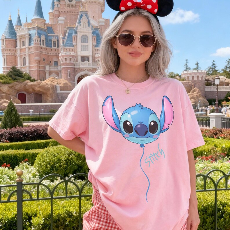 May include: A light pink short-sleeved t-shirt featuring a graphic of the character Stitch from Lilo and Stitch designed as a blue balloon with large ears and a dark nose, with the word Stitch written in blue cursive script below the balloon graphic.