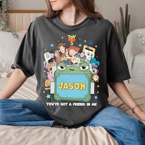 May include: Dark gray t-shirt featuring a colorful graphic of characters from Toy Story 5, including Woody, Buzz Lightyear, and Jessie. A green frog holds a sign that says "JASON". The text "YOU'VE GOT A FRIEND IN ME" is below the frog.