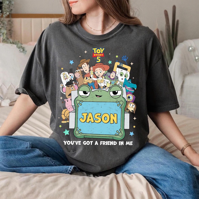 May include: Dark gray t-shirt featuring a colorful graphic of characters from Toy Story 5, including Woody, Buzz Lightyear, and Jessie. A green frog holds a sign that says "JASON". The text "YOU'VE GOT A FRIEND IN ME" is below the frog.