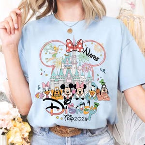 May include: Light blue t-shirt featuring a Disney theme. The design includes the Disney logo, various characters, a castle, fireworks, and the text "Trip 2026". A red bow and the word "Name" are also included.