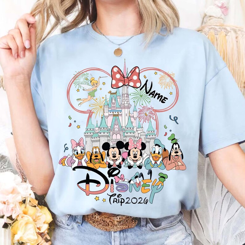 May include: Light blue t-shirt featuring a Disney theme. The design includes the Disney logo, various characters, a castle, fireworks, and the text "Trip 2026". A red bow and the word "Name" are also included.