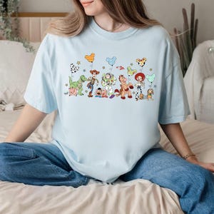 K&ouml;nnte beinhalten: Ein hellblaues T-Shirt mit einem Grafikdruck verschiedener Toy Story-Figuren, darunter Woody, Buzz Lightyear, Jessie, Rex und Slinky Dog. Die Figuren halten bunte, mausohrf&ouml;rmige Luftballons, die von kleinen Sternen umgeben sind.