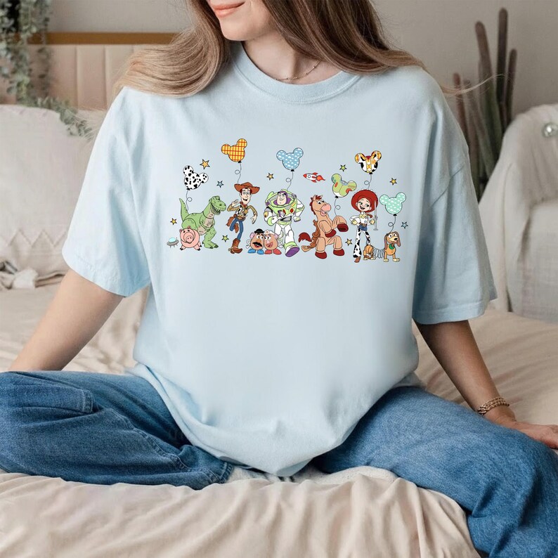 K&ouml;nnte beinhalten: Ein hellblaues T-Shirt mit einem Grafikdruck verschiedener Toy Story-Figuren, darunter Woody, Buzz Lightyear, Jessie, Rex und Slinky Dog. Die Figuren halten bunte, mausohrf&ouml;rmige Luftballons, die von kleinen Sternen umgeben sind.