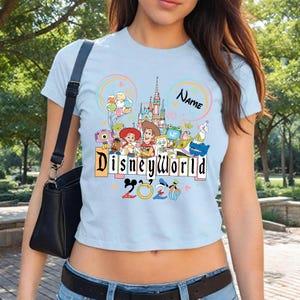 Peut inclure: T-shirt bleu clair avec un motif Disney World. Le motif comprend un ch&acirc;teau, des personnages de dessins anim&eacute;s et l'inscription "Disney World 2020". Un sac noir est visible.