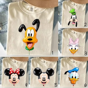 May include: A collection of six cream-colored t-shirts, each featuring a different Disney character designed as a balloon. The characters include Pluto, Goofy, Daisy Duck, Minnie Mouse, Mickey Mouse, and Donald Duck, all with a small green string.