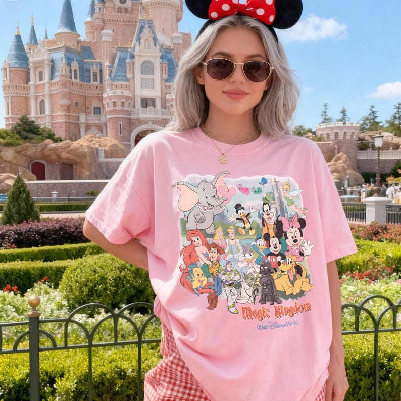 K&ouml;nnte beinhalten: Rosa T-Shirt mit einer Collage von Disney-Figuren, darunter Mickey und Minnie Mouse sowie Dumbo. Auf dem Shirt steht "Magic Kingdom". Die Person tr&auml;gt eine Sonnenbrille und ein Minnie Mouse-Stirnband.