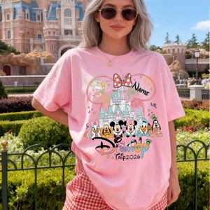 May include: Pink t-shirt featuring a Disney design with Mickey and Minnie Mouse, Donald Duck, Daisy Duck, and Goofy. The design includes a castle, a bow, and the word "Disney". The text "Trip 2026" is also visible.