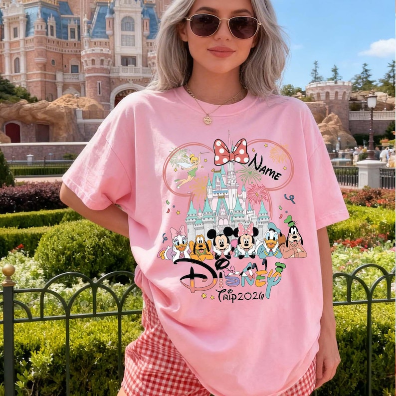 May include: Pink t-shirt featuring a Disney design with Mickey and Minnie Mouse, Donald Duck, Daisy Duck, and Goofy. The design includes a castle, a bow, and the word "Disney". The text "Trip 2026" is also visible.