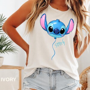 May include: An ivory sleeveless tank top featuring a graphic of a blue Stitch character designed as a round balloon with large pink-lined ears. Below the balloon, the word Stitch is written in a light blue cursive script font on the front.