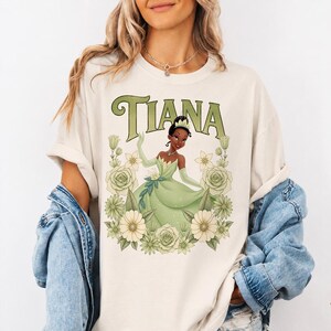 Puede incluir: Camiseta color crema con un gr&aacute;fico de una princesa con un vestido verde, rodeada de flores. La palabra "TIANA" est&aacute; impresa encima de la princesa en letras verdes estilizadas. El dise&ntilde;o est&aacute; centrado en la camiseta.