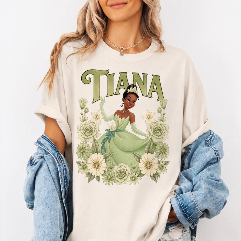 Puede incluir: Camiseta color crema con un gr&aacute;fico de una princesa con un vestido verde, rodeada de flores. La palabra "TIANA" est&aacute; impresa encima de la princesa en letras verdes estilizadas. El dise&ntilde;o est&aacute; centrado en la camiseta.