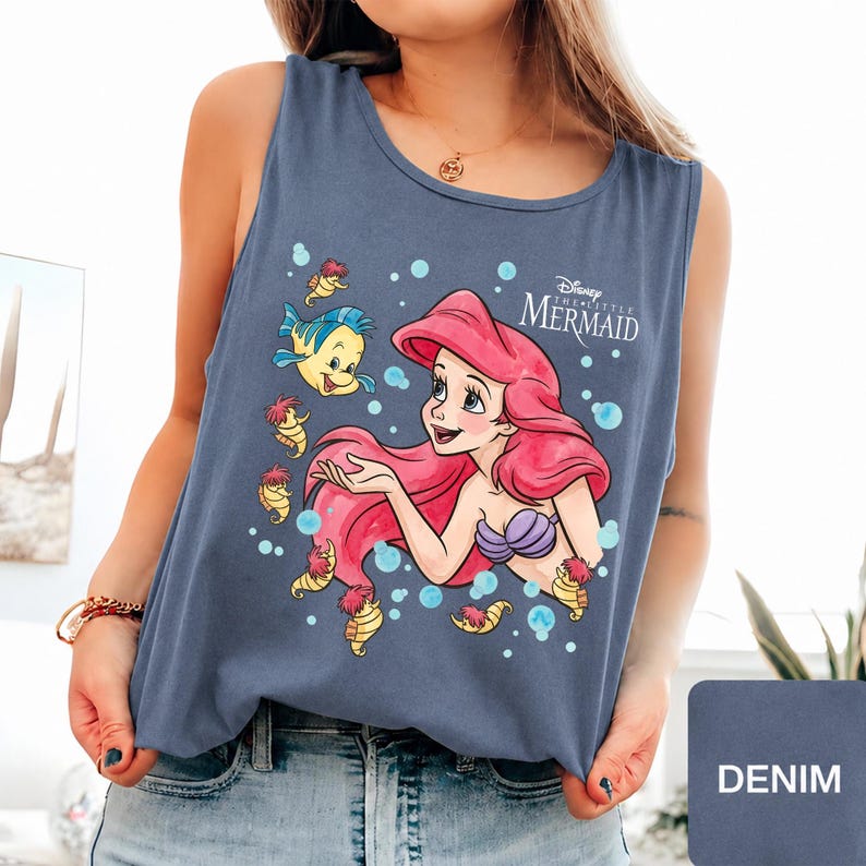 Peut inclure: Un d&eacute;bardeur bleu ardoise sans manches avec une impression graphique d'Ariel de La Petite Sir&egrave;ne aux cheveux rouges, un haut coquillage violet et le poisson Polochon, entour&eacute;s de petits hippocampes jaunes et de bulles bleu clair.