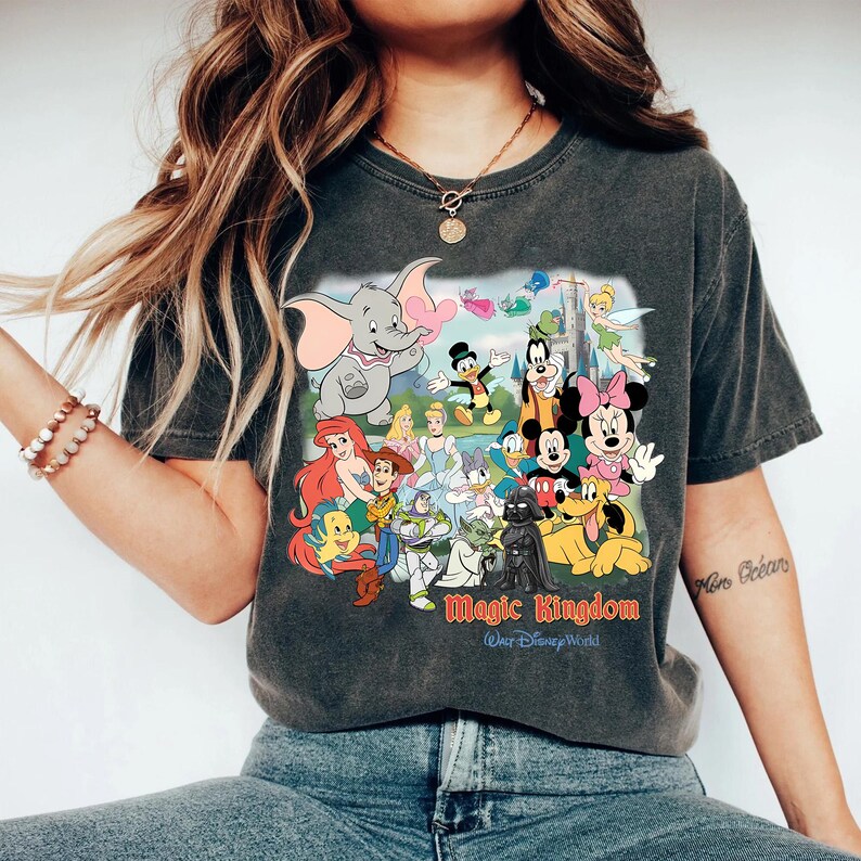 K&ouml;nnte beinhalten: Dunkelgraues T-Shirt mit einer farbenfrohen Grafik von Disney-Figuren wie Dumbo, Mickey Mouse und Arielle. Die Grafik enth&auml;lt den Text "Magic Kingdom Walt Disney World". Das Shirt hat einen Rundhalsausschnitt und kurze &Auml;rmel.