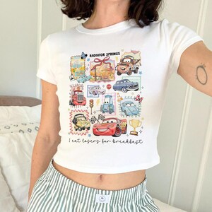 May include: White crop top featuring a colorful graphic of cartoon cars and the text "Radiator Springs" and "I eat lasers for breakfast". The design includes various car illustrations and a trophy.