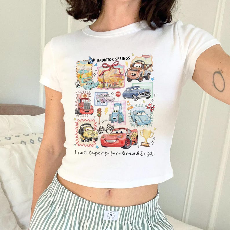 May include: White crop top featuring a colorful graphic of cartoon cars and the text "Radiator Springs" and "I eat lasers for breakfast". The design includes various car illustrations and a trophy.