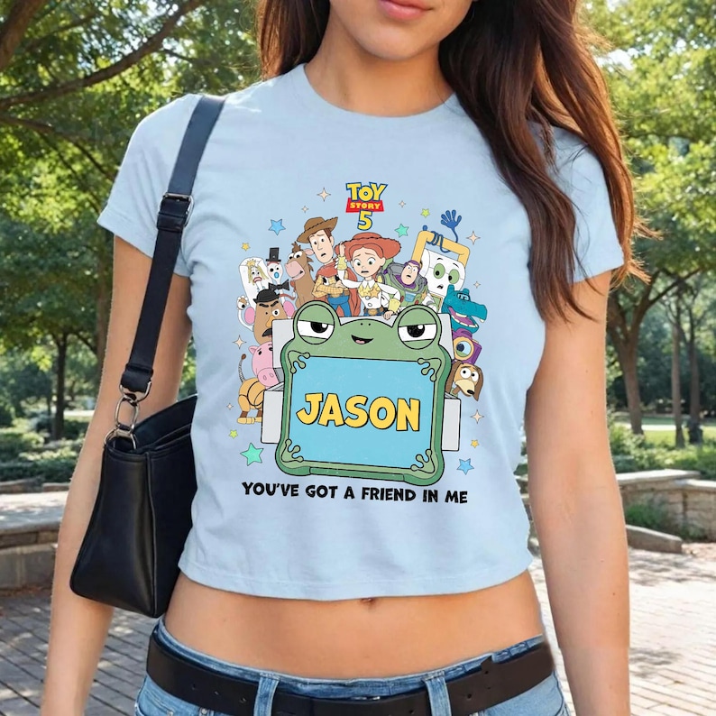 May include: Light blue t-shirt featuring a colorful graphic of characters from Toy Story 5, including Woody and Buzz Lightyear. The shirt has a frog-shaped frame with the name "JASON" and the phrase "YOU'VE GOT A FRIEND IN ME".