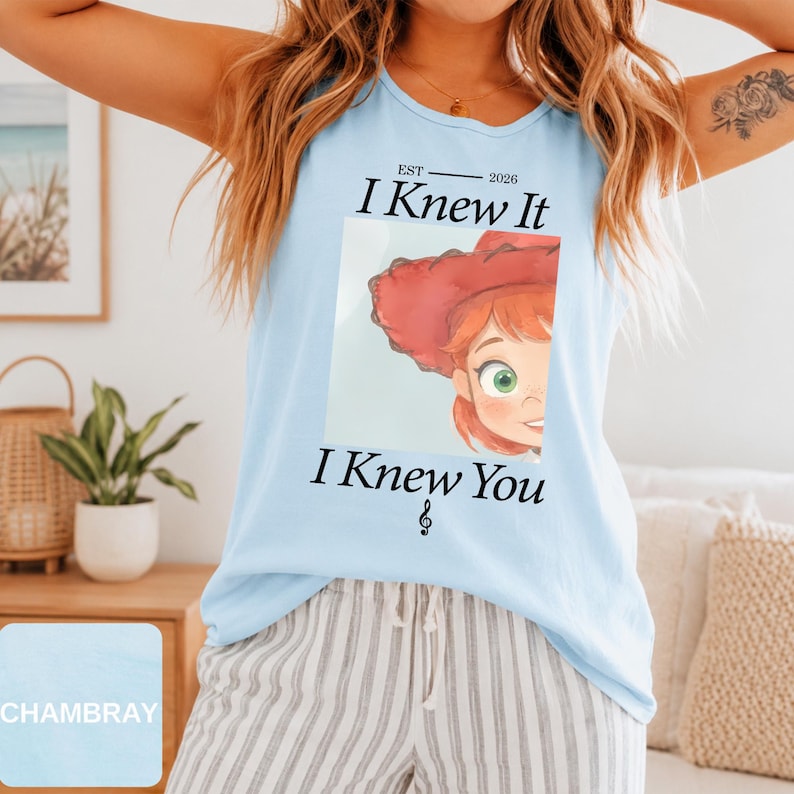 May include: A light blue sleeveless tank top featuring a graphic print of a cartoon character with red hair and a red hat. The text reads EST 2026 I Knew It I Knew You with a small musical note symbol below the text, centered on the chest area.