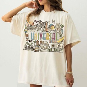 May include: Cream-coloured t-shirt with a colourful graphic of Universal Studios attractions and the text "UNIVERSAL STUDIOS". The design includes cartoon characters, landmarks, and a globe.