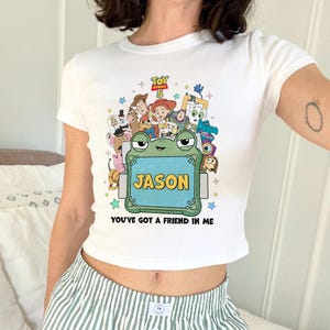 May include: White crop top featuring a colorful graphic of Toy Story characters, the text "TOY STORY 5", and the name "JASON" on a frog-shaped sign. The phrase "YOU'VE GOT A FRIEND IN ME" is also printed on the shirt.