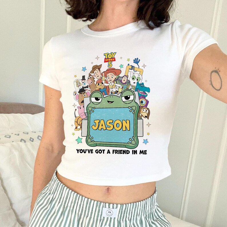 May include: White crop top featuring a colorful graphic of Toy Story characters, the text "TOY STORY 5", and the name "JASON" on a frog-shaped sign. The phrase "YOU'VE GOT A FRIEND IN ME" is also printed on the shirt.