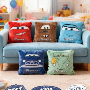 May include: Five plush throw pillows featuring cartoon car designs in red, brown, blue, dark blue, and green. Each pillow has a smiling car face and the name "Thomas" embroidered on it. The pillows are arranged on a light blue couch.