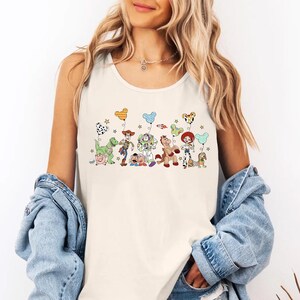May include: Cream-colored tank top featuring a colorful graphic of Toy Story characters holding balloon bouquets. The design includes Woody, Buzz Lightyear, and other characters. The balloons have Mickey Mouse, Dalmatian, and other patterns.