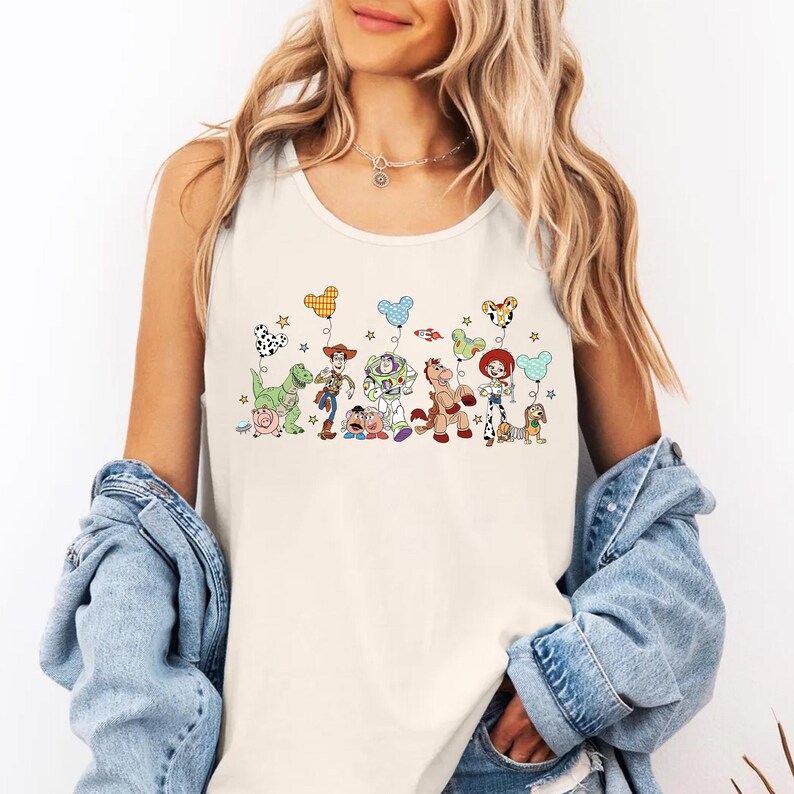 May include: Cream-colored tank top featuring a colorful graphic of Toy Story characters holding balloon bouquets. The design includes Woody, Buzz Lightyear, and other characters. The balloons have Mickey Mouse, Dalmatian, and other patterns.