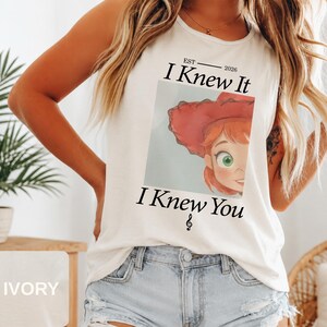 May include: An ivory sleeveless tank top featuring a graphic print of a cartoon character with red hair and a red hat. The text reads EST 2026 I Knew It I Knew You with a small musical note symbol below the text, centered on the front of the shirt.