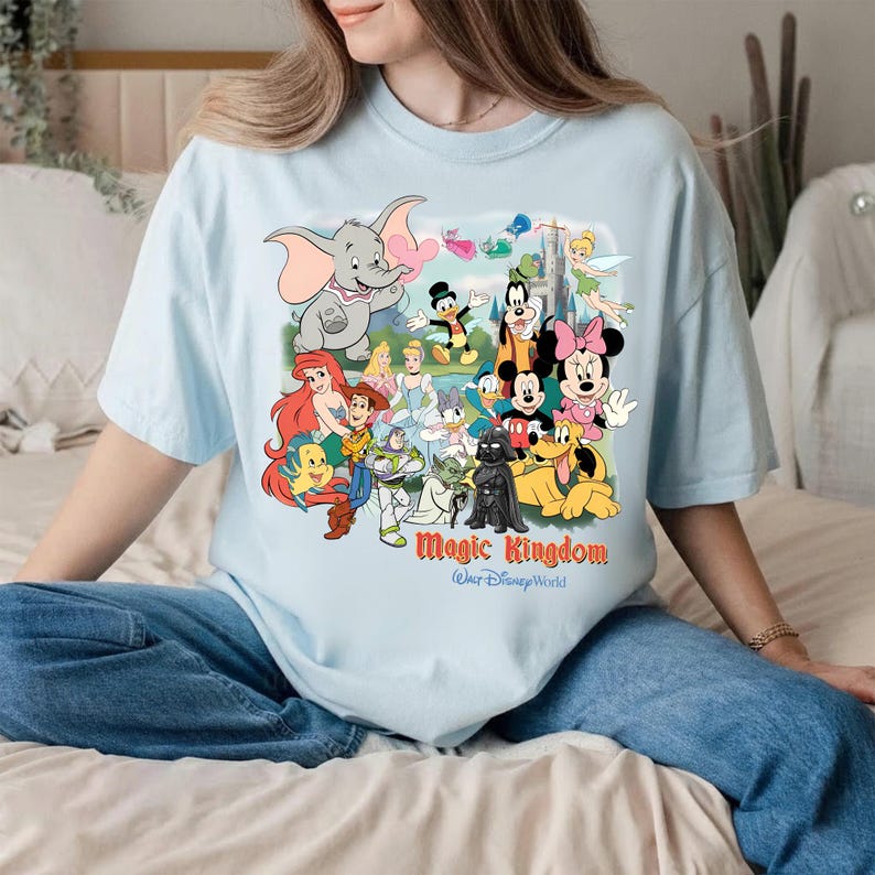 K&ouml;nnte beinhalten: Hellblaues T-Shirt mit einem farbenfrohen Aufdruck von Disney-Figuren, darunter Mickey Mouse und Dumbo. Der Text "Magic Kingdom Walt Disney World" ist in Rot und Gold gedruckt. Das Shirt ist aus weichem Material.