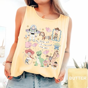 May include: A pale yellow sleeveless tank top featuring a graphic print of various animated characters including a space ranger, a cowboy, a cowgirl, a dinosaur, a pig, and a dog. The text "YOU'VE GOT A FRIEND IN ME" is printed in the center.